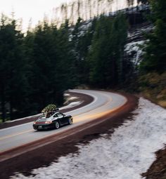 a car driving down a winding road with trees in the background and snow on the ground