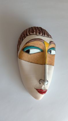 a white mask with blue, yellow and brown designs on it's face is hanging from the wall