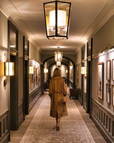 a woman walking down a long hallway with lights on either side of her and pictures on the wall