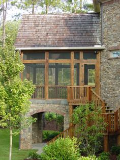 a stone house with a wooden porch and stairs leading up to the front door is surrounded by greenery