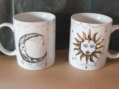 two coffee mugs sitting on top of a table next to each other, one with a sun and moon painted on it