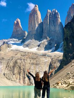 two women standing on the edge of a lake with their arms outstretched in front of mountains