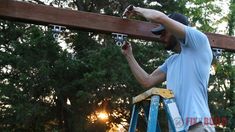 a man standing on a ladder working on a wooden beam in front of some trees