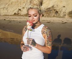 a woman with tattoos holding a flower in her hand and looking at the camera while standing on the beach