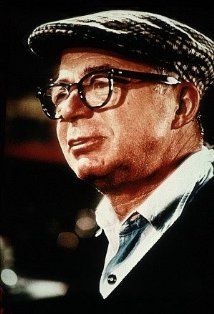 a man wearing glasses and a hat looks off into the distance while standing in front of an audience