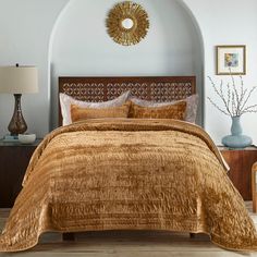 a bed with gold comforter and pillows in a white walled room next to a wooden headboard