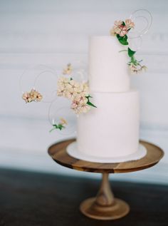 a three tiered white wedding cake with flowers on the top and bottom, sitting on a wooden stand