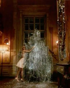 a woman standing next to a giant christmas tree in front of a window with lights on it