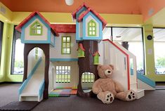 a teddy bear is sitting in front of a toy house with stairs and trees on the floor