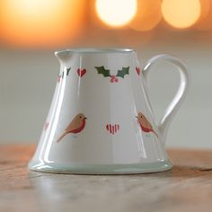 a white pitcher with red and green designs on it sitting on a wooden table in front of some lights