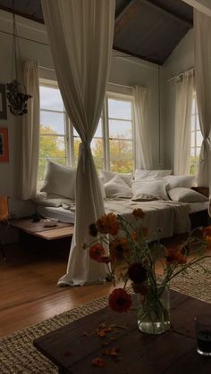 a bedroom with white curtains and flowers in the vase on the coffee table next to the bed