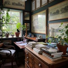 a desk with many books and plants on it in front of two large windows filled with framed pictures