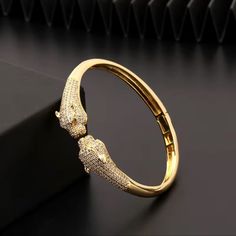 This Bracelet Is A Luxurious Gold Bangle Featuring Two Intricately Designed Panther Heads Facing Each Other At The Center. The Panthers Are Meticulously Detailed, With Sparkling Stones Encrusted Across Their Bodies, Adding A Touch Of Elegance And Brilliance To The Piece. Their Eyes Are Accentuated, Possibly With Colored Stones Or Gems, Which Adds Depth To The Animal Motif. The Bracelet Has A Sleek, Polished Gold Finish, And The Hinged Clasp Ensures A Seamless, Secure Fit. The Combination Of The Powerful Panther Design And The Dazzling Stones Make This A Statement Accessory, Symbolizing Strength, Grace, And Sophistication. This Type Of Design Is Often Favored For Its Bold Yet Timeless Aesth Panther Design, Anthropologie Jewelry, Colored Stones, Gold Bangle, High End Fashion, Gold Bangles, Bangle Bracelet, Gold Finish, Panther