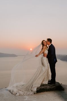 a bride and groom standing on top of a cliff at sunset with veil blowing in the wind