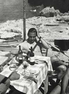 a man sitting at a table with two bottles of wine in front of him on the beach