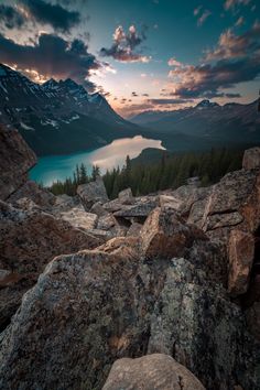 the sun is setting over a lake surrounded by rocks and pine trees with mountains in the background