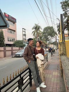 two people standing on the side of a road next to a fence and buildings with power lines above them