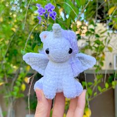 a hand holding a small stuffed animal in front of some trees and bushes with purple flowers