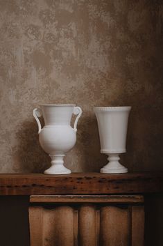 two white vases sitting on top of a wooden table next to each other in front of a wall