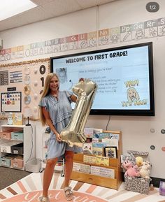 a woman holding up a giant balloon in front of a large tv screen with the words welcome to first grade written on it