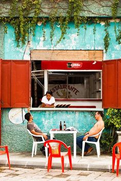 two men sitting at a table in front of a blue building with red shutters