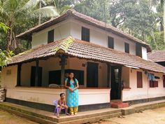 two people standing in front of a house with trees behind them and one person sitting on the porch