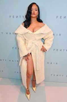 a woman in a white coat and high heels standing next to a wall with the words fenty hair written on it