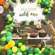 a giraffe standing next to a table filled with cakes and desserts in front of a sign that says mason wild one