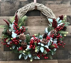 a wreath with berries, pine cones and greenery is hanging on a wooden wall