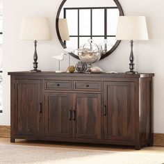 a wooden sideboard with two lamps and a round mirror on the wall behind it