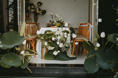 a dining room table with white flowers and greenery on it, surrounded by wooden chairs