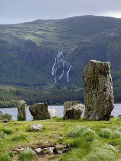 some large rocks in the grass near water and mountains with a waterfall coming out of it