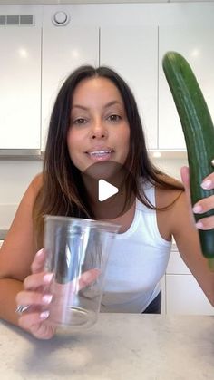 a woman holding a cucumber and glass in her hand while sitting at a kitchen counter