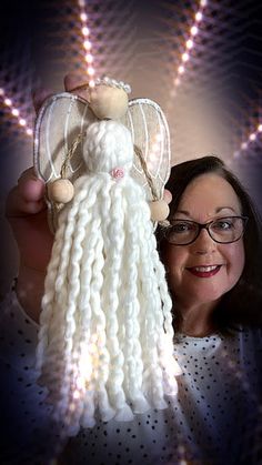 a woman holding up a white knitted angel ornament with lights in the background