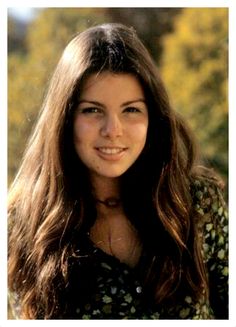 a woman with long brown hair smiling at the camera and wearing a floral shirt in front of trees