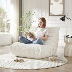 a woman sitting on a white chair reading a book in her living room with a potted plant