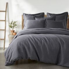 a bed with grey linens and pillows in a white room next to a ladder