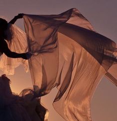 a woman in a white dress is holding a sheer fabric over her head as the sun sets behind her