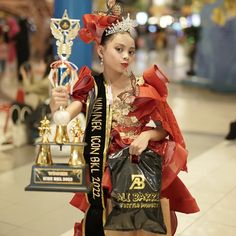 a woman in a costume holding two trophies and wearing a tiara with red ribbons