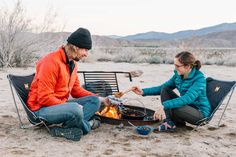 a man and woman sitting next to each other on top of a sandy beach near a campfire