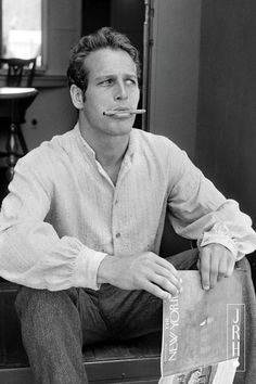 black and white photograph of man sitting on bench with newspaper in mouth, looking at camera