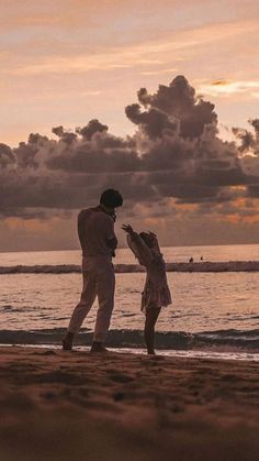 a man and woman standing on top of a beach next to the ocean at sunset