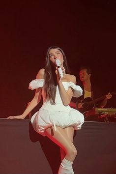 a woman in white dress on stage holding a microphone and singing into a microphone with two men standing behind her