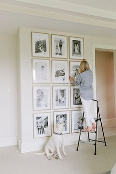 a woman standing on a ladder next to a wall with pictures and a white dog