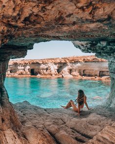 a woman sitting on the rocks looking out at the ocean from inside a cave with blue water