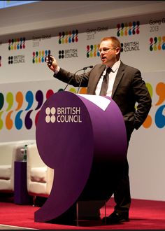 a man in a suit and tie standing at a podium with his cell phone up to his ear