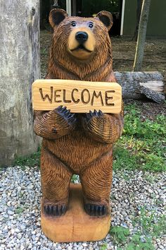 a bear statue holding a welcome sign in front of a wooden post with the word welcome written on it