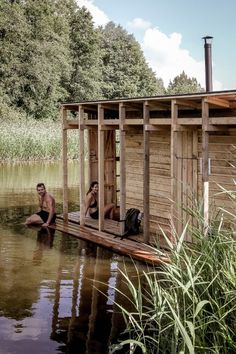 two people are sitting in the water near a small wooden cabin on stilts that is surrounded by tall grass and trees