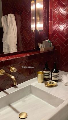 a white sink sitting under a bathroom mirror next to a red tiled wall and gold faucet