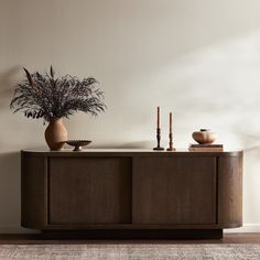 a vase with some plants on top of it next to a wooden cabinet and two candles